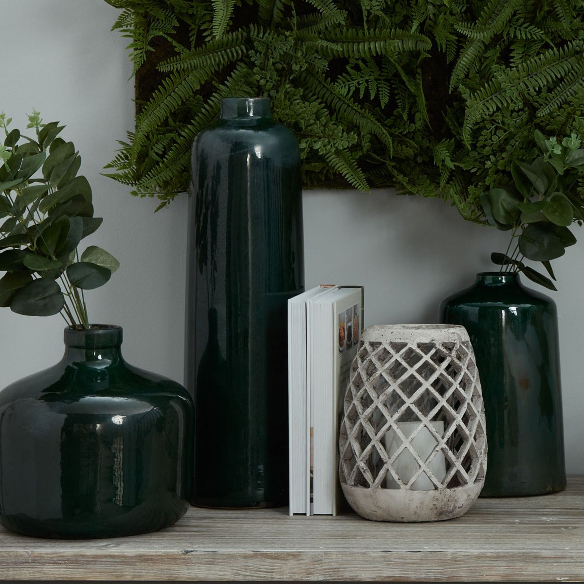 Emerald Eve Vase at left with foliage, shown alongside taller green vases, books and a lattice candle holder