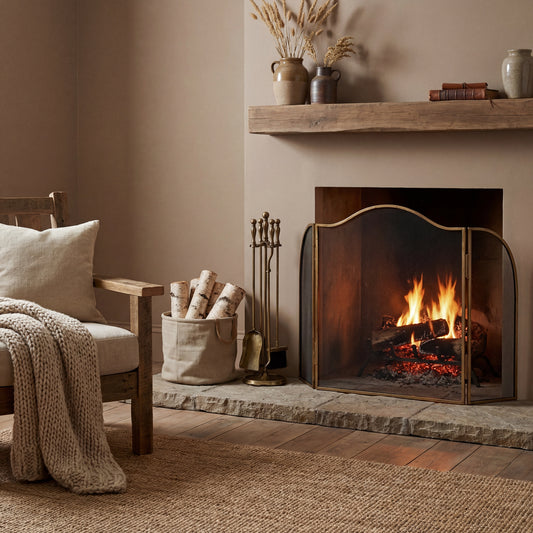 Antiqued brass large fire screen with arched mesh panels in front of a lit fireplace in a warm neutral living room — Xquisite By Design
